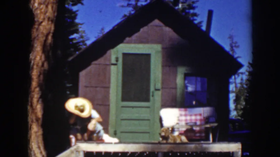 Late-1930s Lake Tahoe home-movie frame showing a straw-hatted person leaning on a rustic cabin porch near a green door.