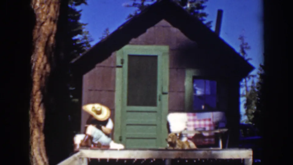 In 1939 Lake Tahoe home-movie footage, a woman in a wide straw hat sits outside a small cabin while a dog rests near the porch.
