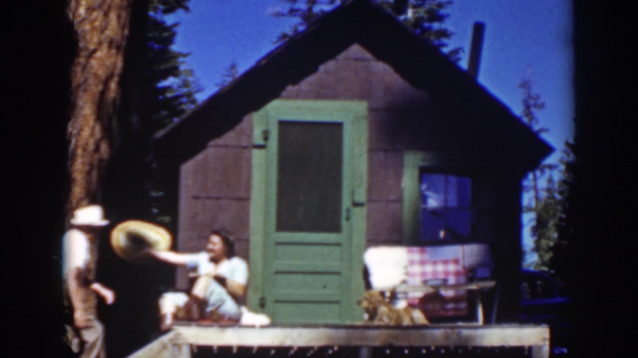 Man approaches a seated woman outside a small cabin as a dog stays near the door.