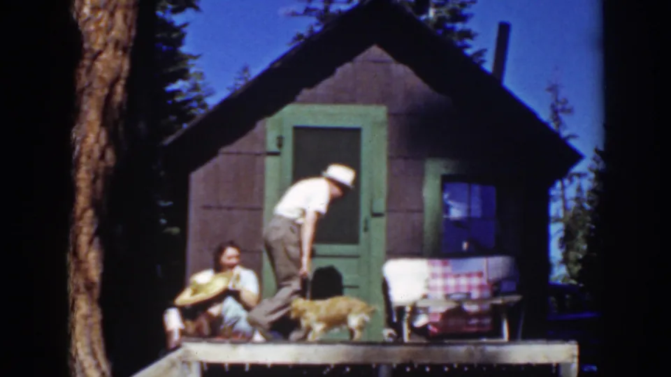 A man stands on the porch beside the seated woman and dog in front of a small cabin with green trim in Lake Tahoe footage.