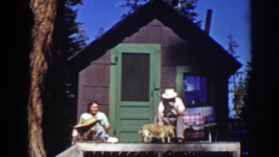 Lake Tahoe 1939 home-movie frame with a woman holding a straw hat and a man leaning forward beside a dog on a cabin porch.