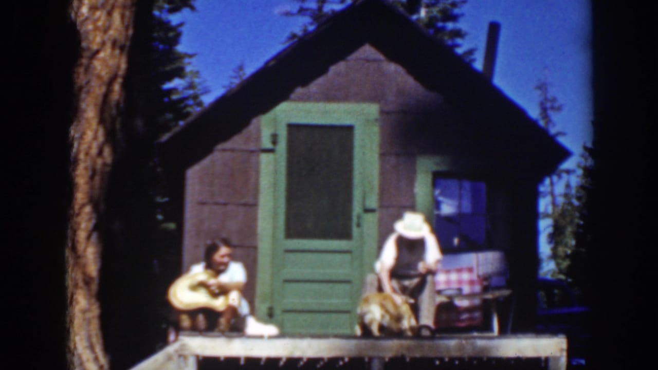 Two adults sit on a cabin porch with a small dog near a green door.