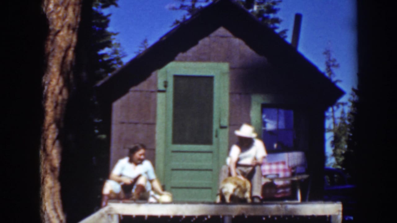 A seated couple pets a small dog on a porch outside a cabin.