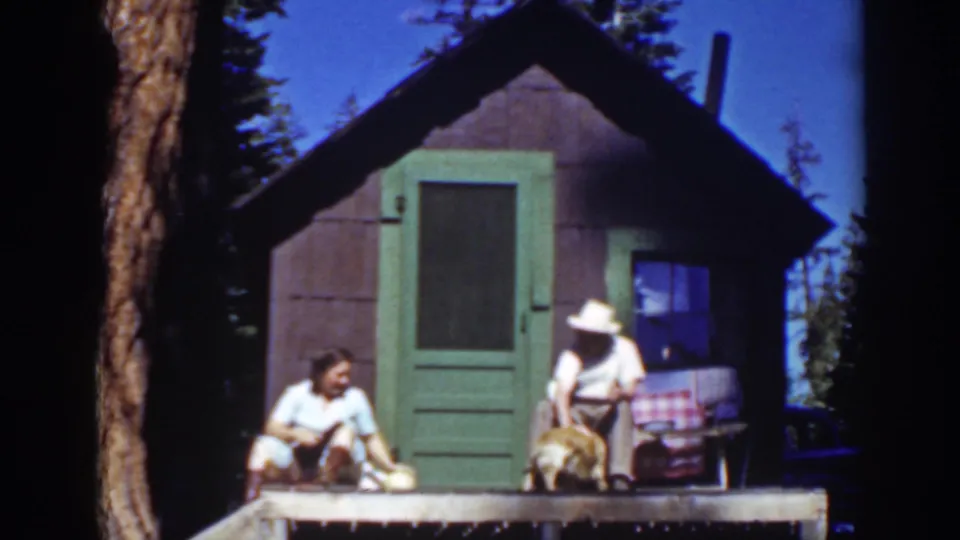The couple remains on the Lake Tahoe cabin porch, with the woman seated at left and the hat-wearing man leaning toward the dog.