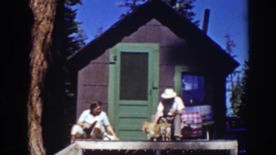 A 1939 Lake Tahoe cabin scene shows the woman seated at left and the man in a white hat bending toward the small dog near the doorway.