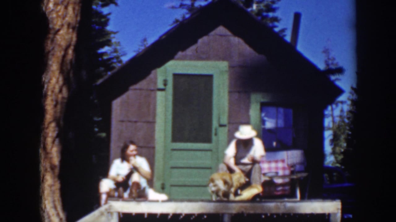 Two adults sit on a small cabin porch with a dog between them.