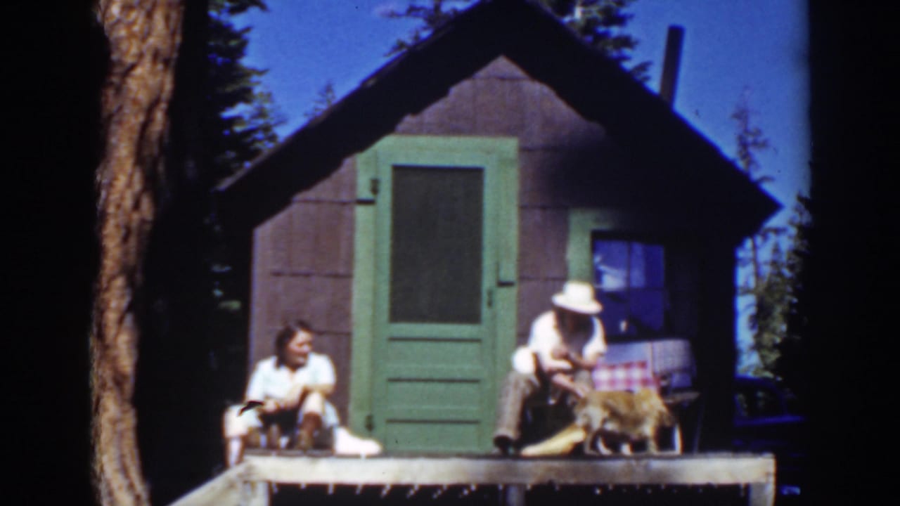 A man in a white hat leans toward a dog while a woman sits nearby on the porch.