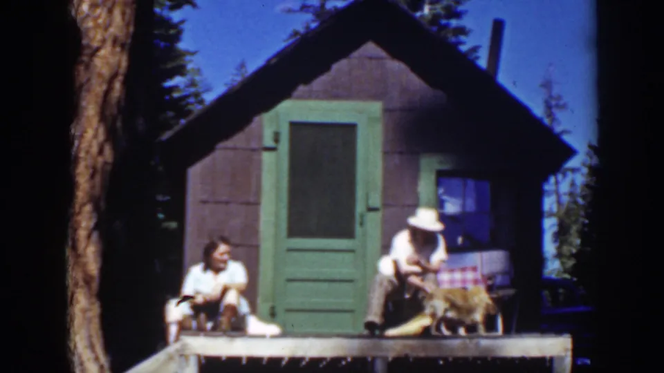 At a Lake Tahoe cabin porch in 1939, a man in a white hat bends toward a dog as a woman sits to the left.