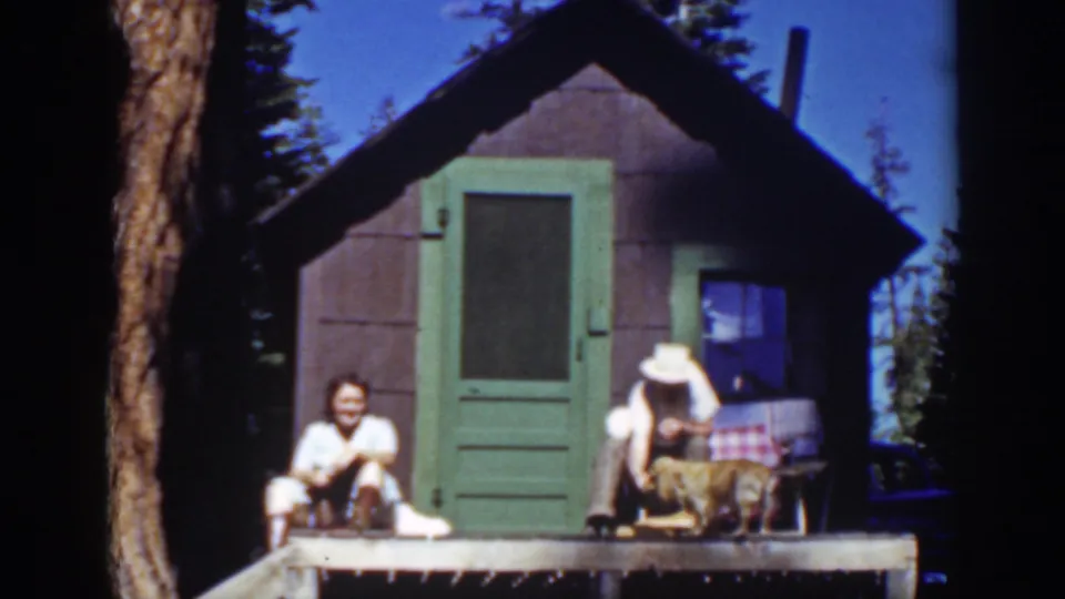 1939 home-movie frame of a woman facing the camera and a man tending to a dog outside a small Lake Tahoe cabin.