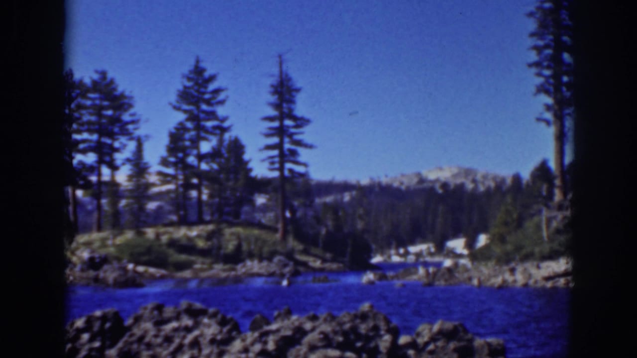 Blue mountain lake bordered by rocks, pine trees, and distant snow-capped peaks.