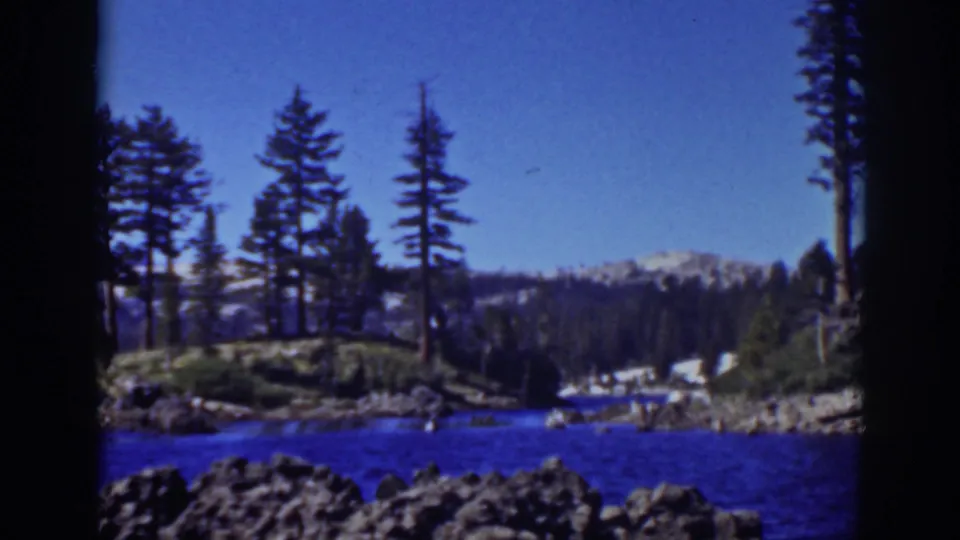 1939 Lake Tahoe home-movie frame showing blue water, rocky foreground, tall pines, forested slopes, and snow patches on distant peaks.