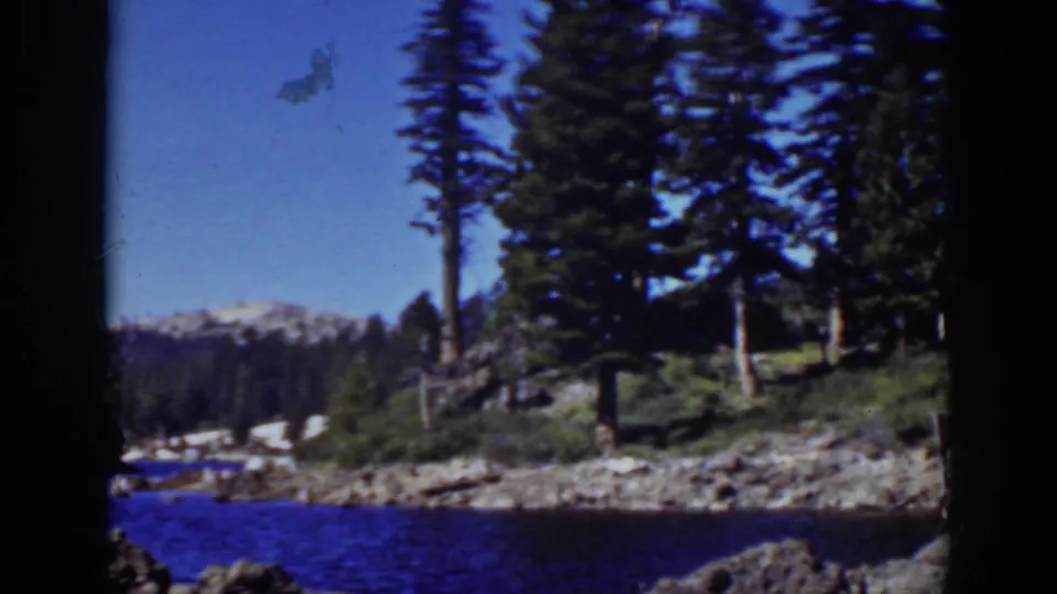 1939 Lake Tahoe frame looking past blue water toward a rocky pine shoreline and snow-streaked mountains in the background.