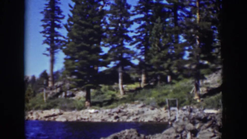 Lake Tahoe 1939 8mm frame showing a rocky pine shoreline beside blue water, with a small dock-like structure near the right edge.