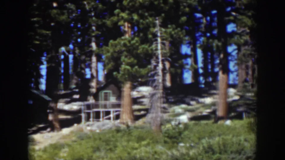 A 1939 Tahoe woodland frame with the cabin partly leaving view as the camera pans across pines, rocks, and low vegetation.