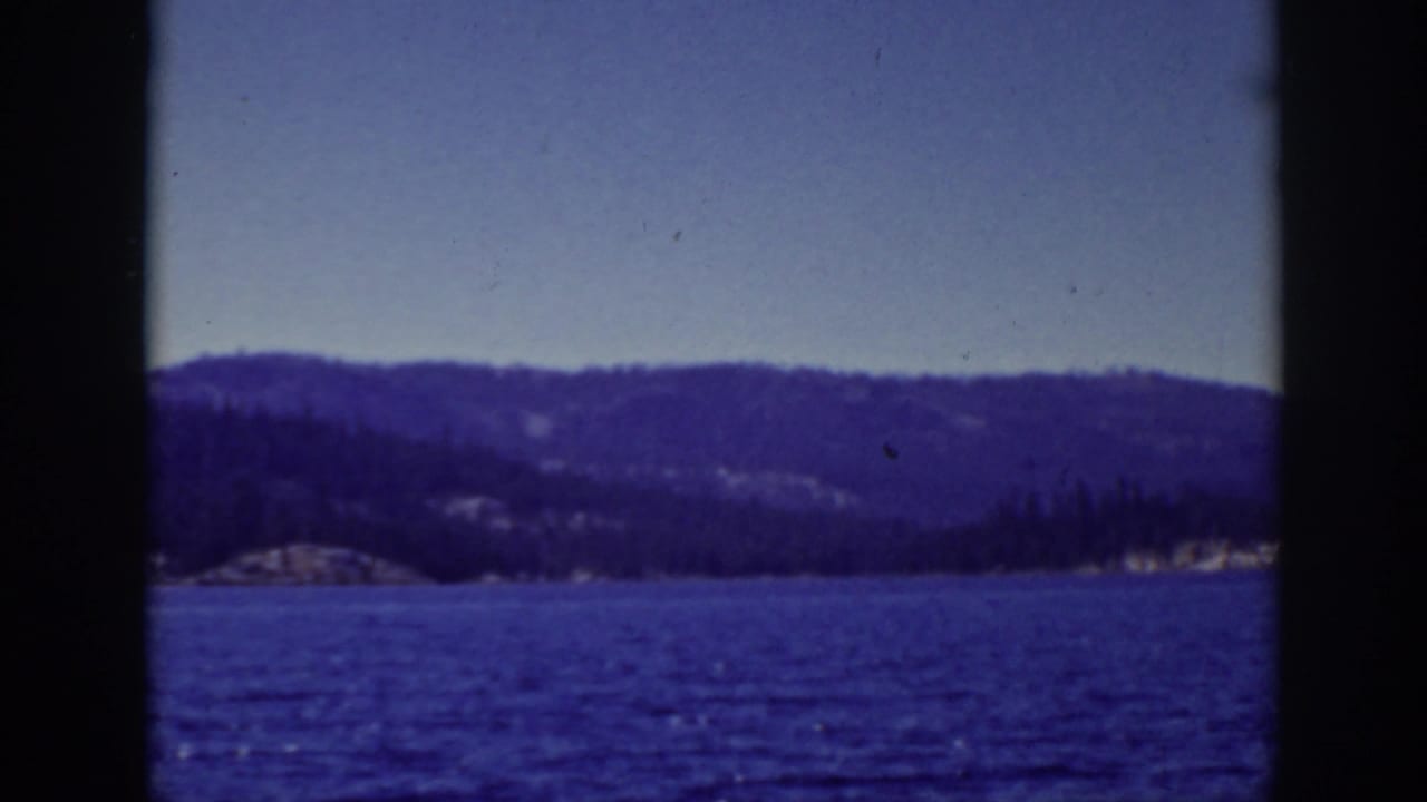 1939 Lake Tahoe frame looking across blue water toward dark wooded hills and a pale clear sky.