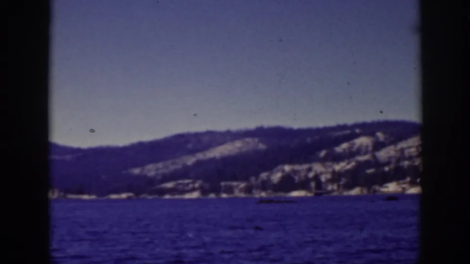 1939 Lake Tahoe frame showing blue water below a distant shoreline and snow-dusted mountain slopes, softened by home-movie focus.