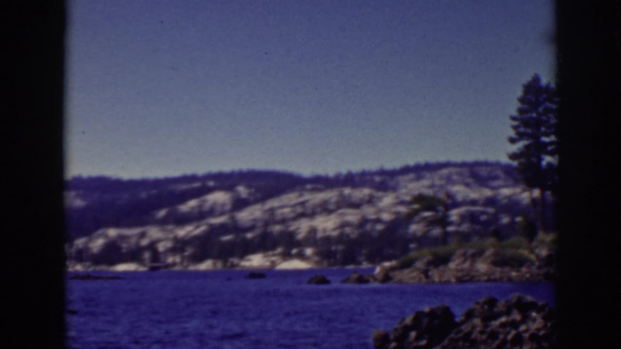 Lake Tahoe scene with blue water, snow-dusted hills, rocky shoreline, and a cluster of pine trees along the right side.