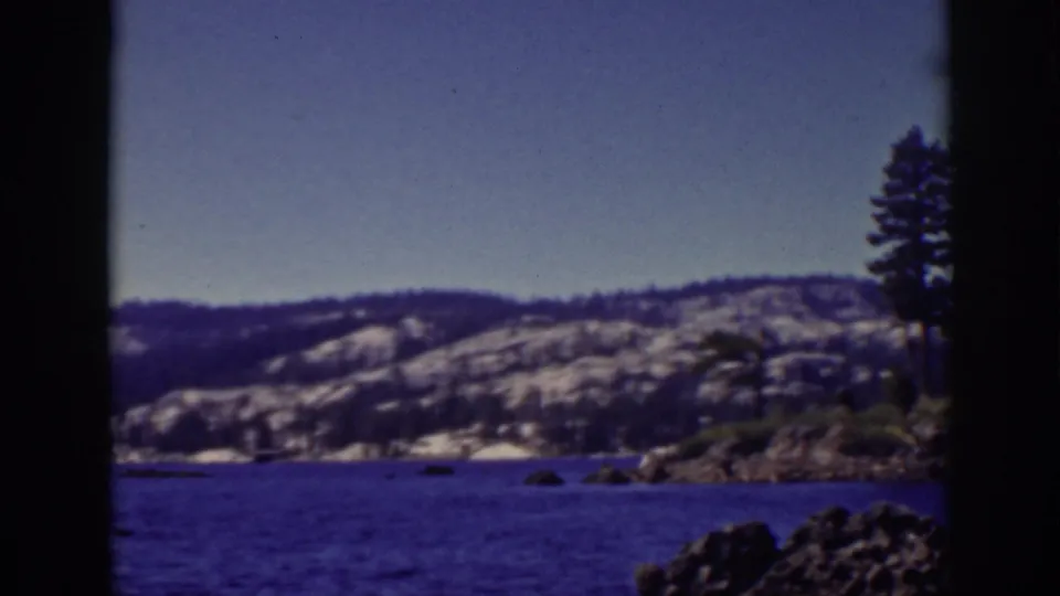 Lake Tahoe scene with blue water, snow-dusted hills, rocky shoreline, and a cluster of pine trees along the right side.