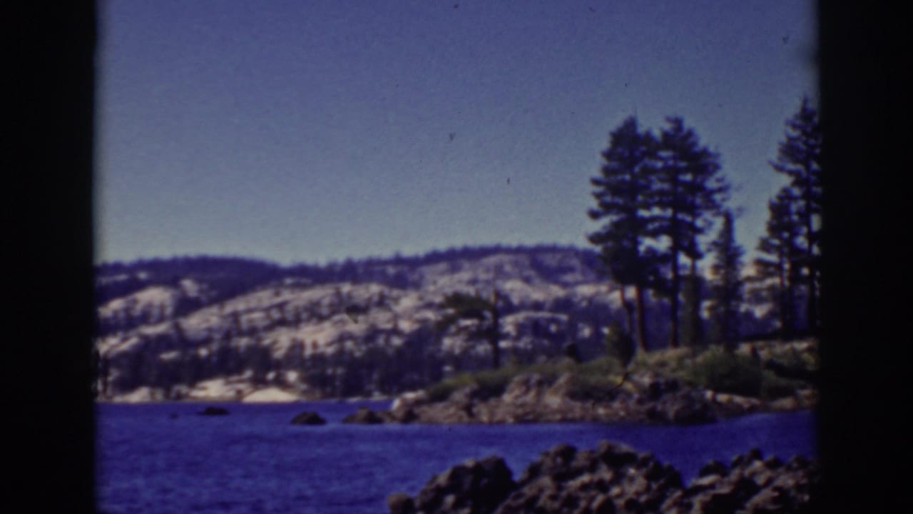1939 Lake Tahoe frame showing a rocky foreground, pine trees on shore, blue water, and snow-dusted hills beyond.