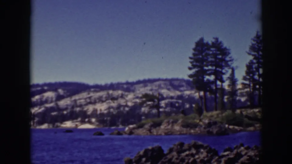 1939 Lake Tahoe frame showing a rocky foreground, pine trees on shore, blue water, and snow-dusted hills beyond.