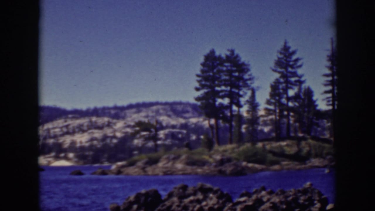 Lake Tahoe home-movie view of pines on a rocky point, blue water, distant snow-dusted hills, and dark 8mm frame edges.