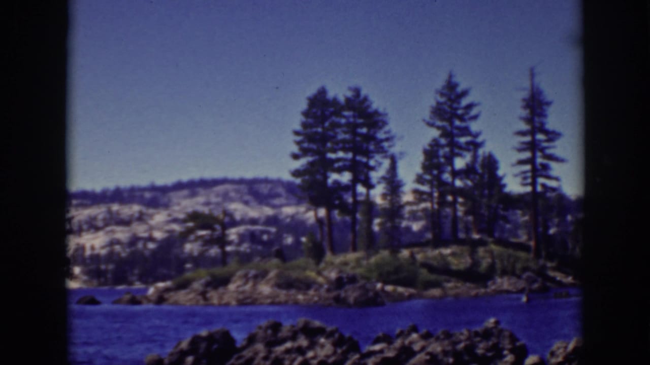 Rocky Lake Tahoe shoreline with blue water, pine trees, hills, and clear sky.