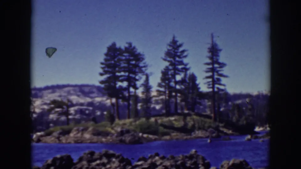 1939 Lake Tahoe home-movie frame showing a pine-covered lakeside point, blue water, rocky shore, distant hills, and clear sky.