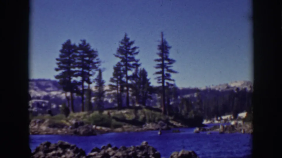 1939 Lake Tahoe scene with blue water, rocks in the foreground, tall pines on shore, forested hills, and distant snow patches.