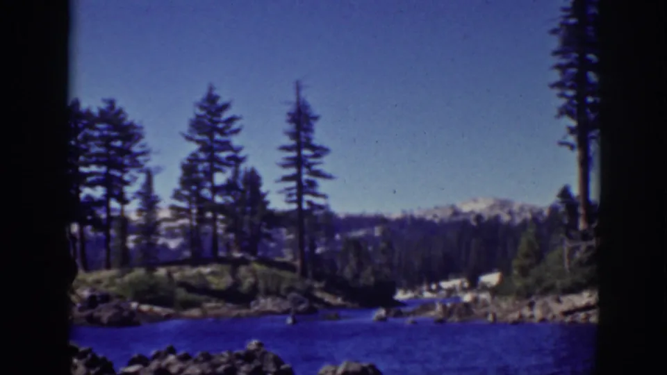 1939 Lake Tahoe frame with calm blue water, rocky shoreline, tall pines, dark forested hills, distant snow, and clear sky.