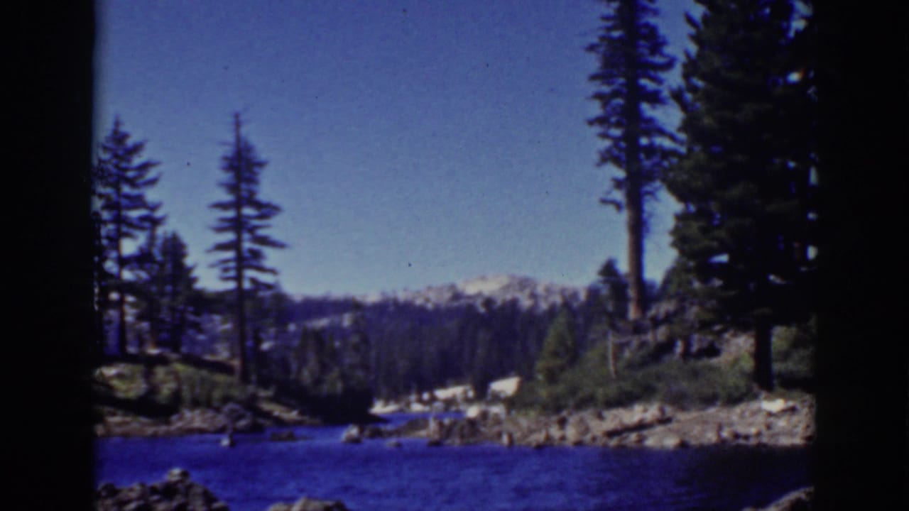 Wide lake view with evergreen trees, rocky shore, distant mountains, and blue sky.