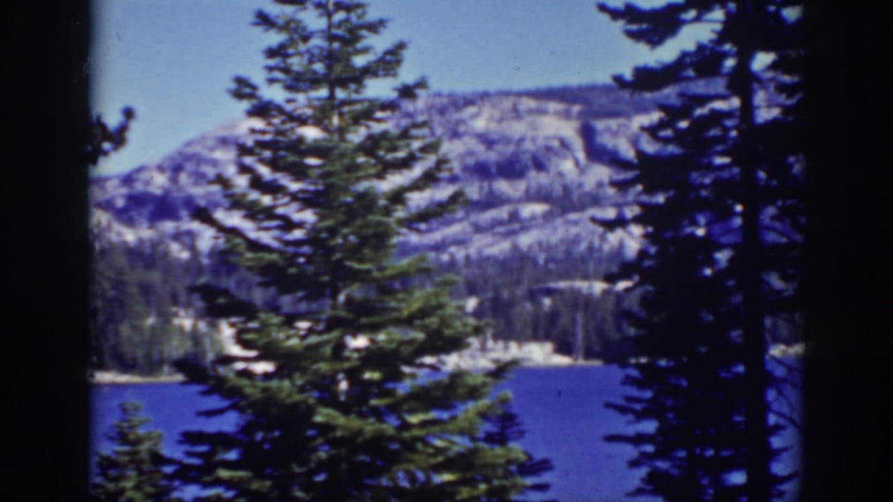 Pine trees frame a blue lake with snow-dusted mountains in the distance.