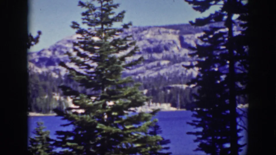 Static 8mm view of Lake Tahoe with pine silhouettes, blue water, and distant snow-streaked mountains.