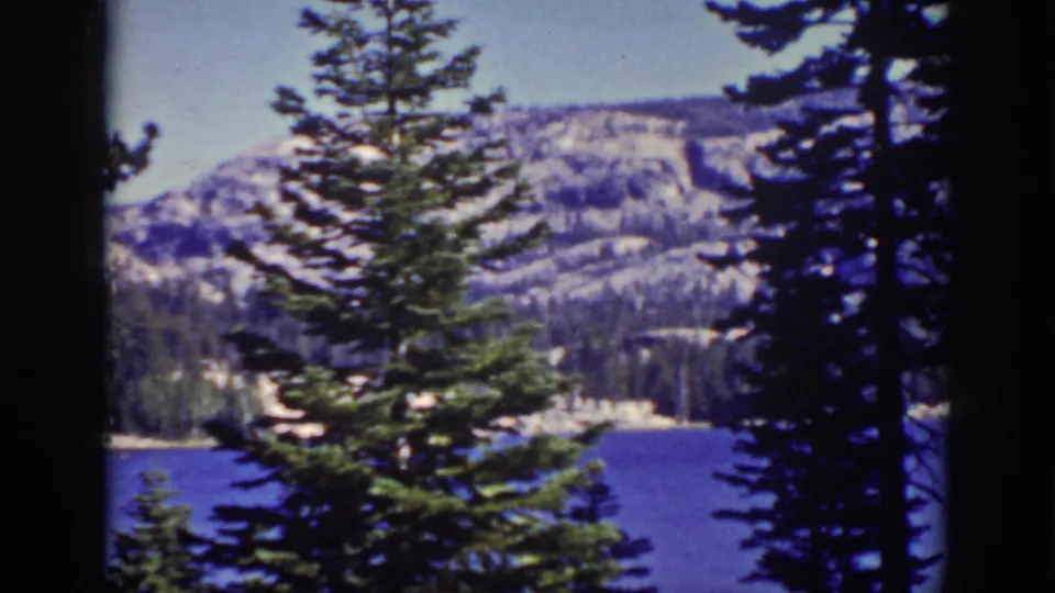Final Lake Tahoe frame from a 1939 8mm home movie, with pine trees framing blue water, shoreline forest, and snow-marked mountains.
