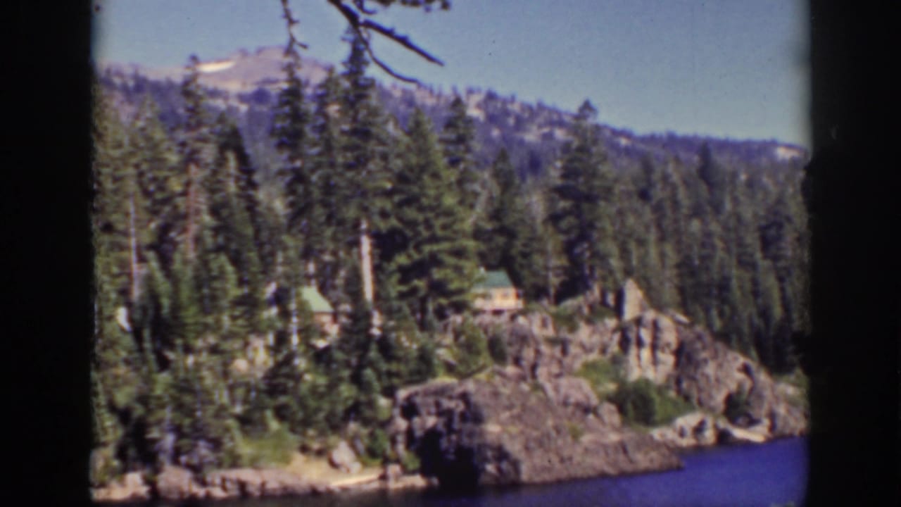 Rocky forested shoreline with small cabins beside blue lake water.