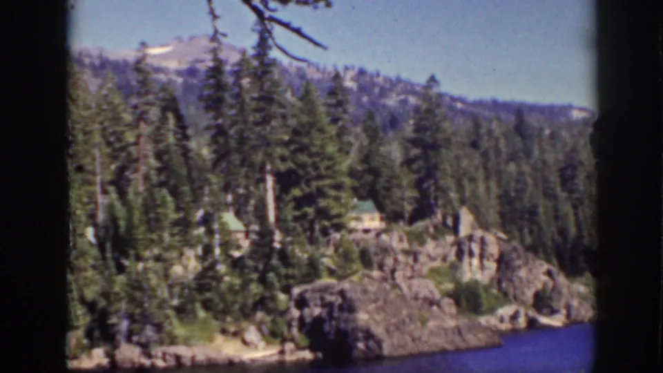 1939 Lake Tahoe frame showing dense pines, rocky lakeside terrain, small cabins, and distant mountain slopes.