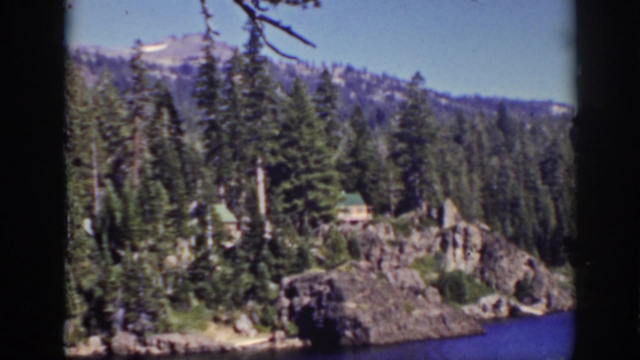 Pine forest, rocky lakeshore, small green-roof cabin, blue water, and distant mountains.