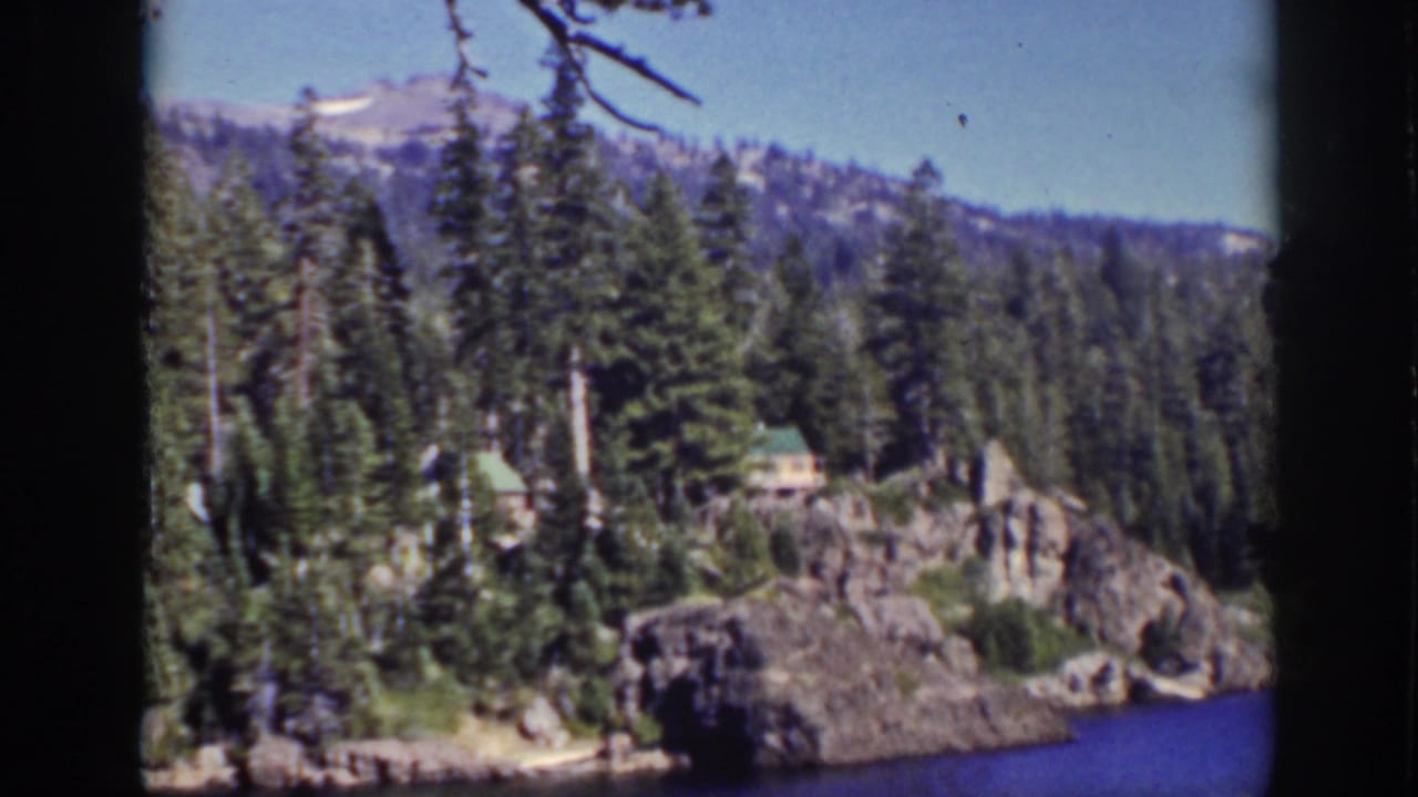 Rocky pine-covered lakeshore with cabin roofs, blue water, wooded hills, and pale sky.