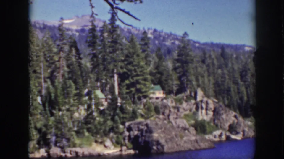 1939 Lake Tahoe view of pine-covered rocks, small lakeside cabins, blue water, wooded hills, and a pale mountain sky.