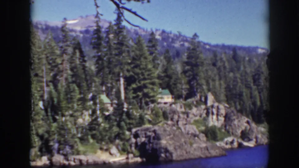 1939 Lake Tahoe 8mm view of a green-roof cabin tucked among pines on rocky terrain beside vivid blue lake water.