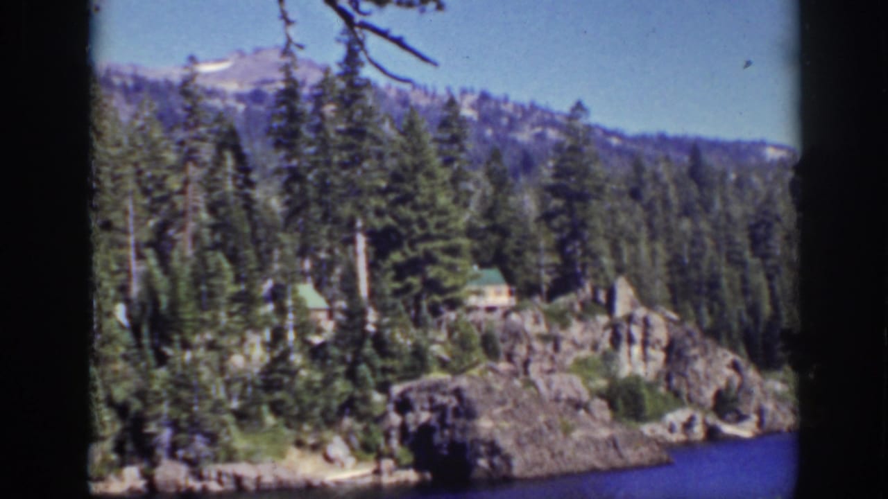 Evergreen-lined Lake Tahoe shore with a small cabin and distant mountains.
