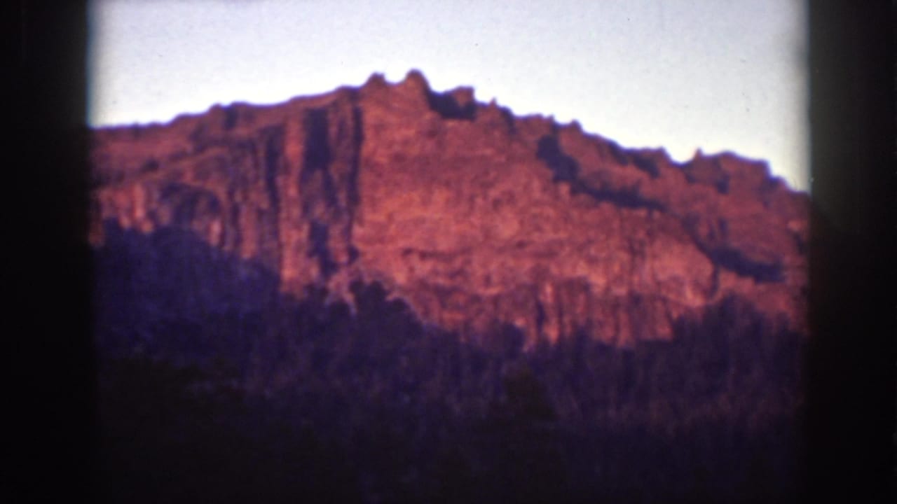 1939 Lake Tahoe 8mm frame showing a warm reddish rocky ridge rising above dark evergreen forest beneath a pale sky.