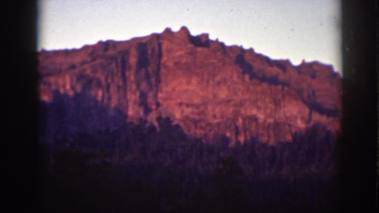 Sunlit reddish mountain cliff above a dark forested slope and pale sky.