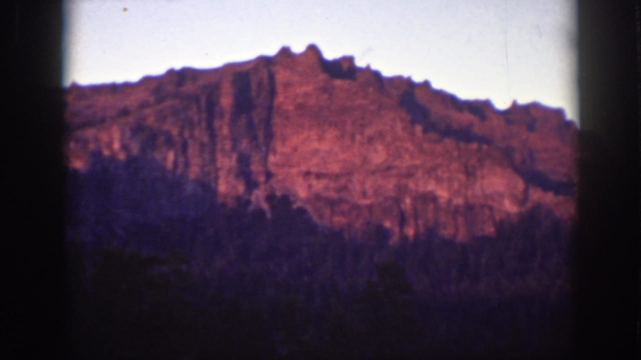 Reddish mountain cliff above a dark tree line with soft sky and dark film edges.