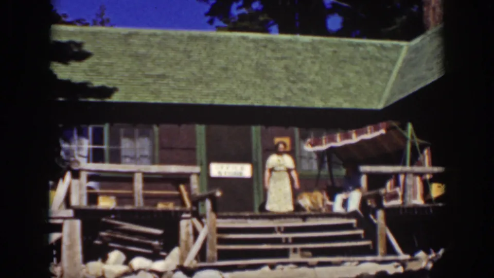 1939 Lake Tahoe frame with a woman in a light dress standing on the steps of a rustic cottage porch.