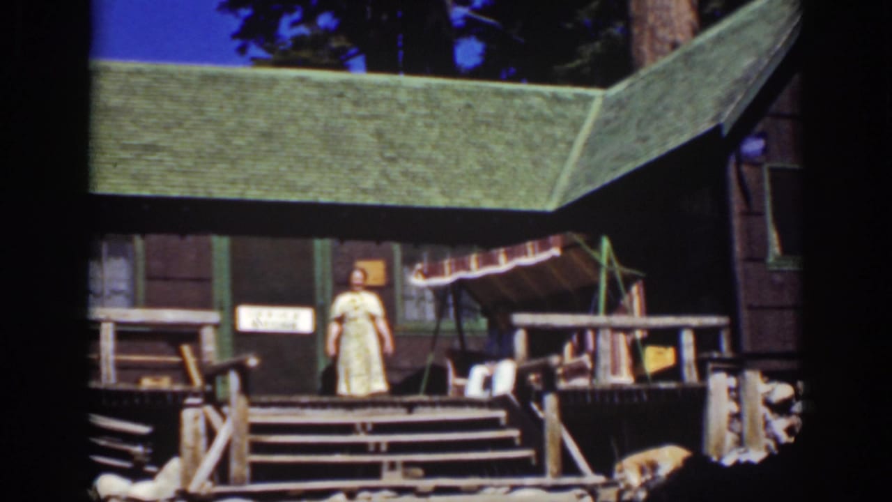 Rustic cabin porch with a woman standing near the doorway and a seated person beside the railing.