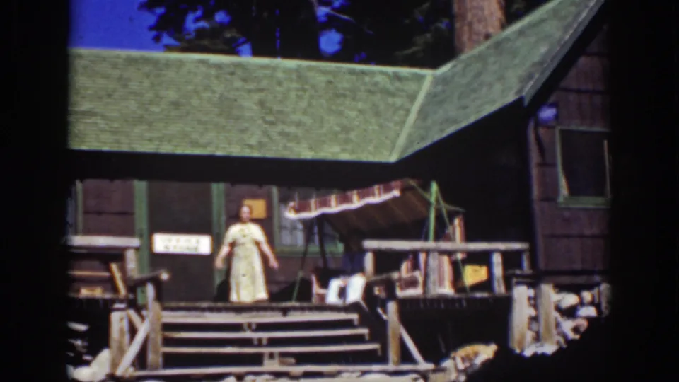 1939 Lake Tahoe frame showing a woman posed on the steps of a rustic wooden cabin with an awning and railings.