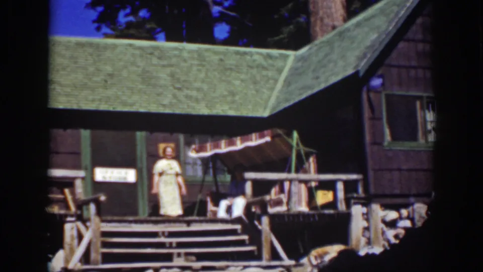 Lake Tahoe vacation-cabin frame with a standing woman, partly visible seated person, porch rails, awning, and green roof.