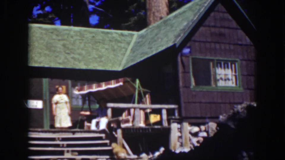 1939 Lake Tahoe home-movie still of a woman standing outside a rustic cabin with porch steps and an awning visible.