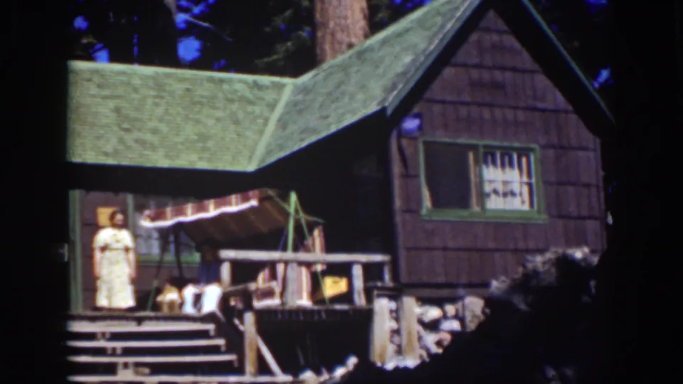 1939 Lake Tahoe frame of a posed woman on a rustic cabin porch, with roof shingles, awning, railings, and trees visible.
