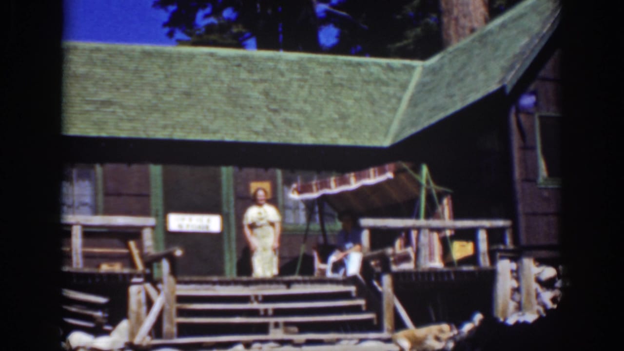 Woman in a light dress stands on porch steps beside a rustic cabin and striped awning.